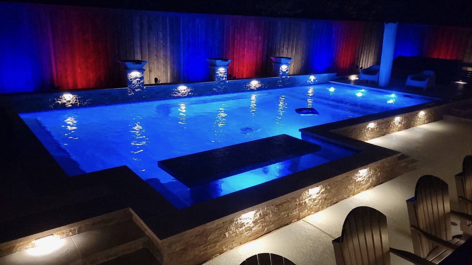 Modern backyard pool illuminated with red, white, and blue LED lighting and stone accents at night
